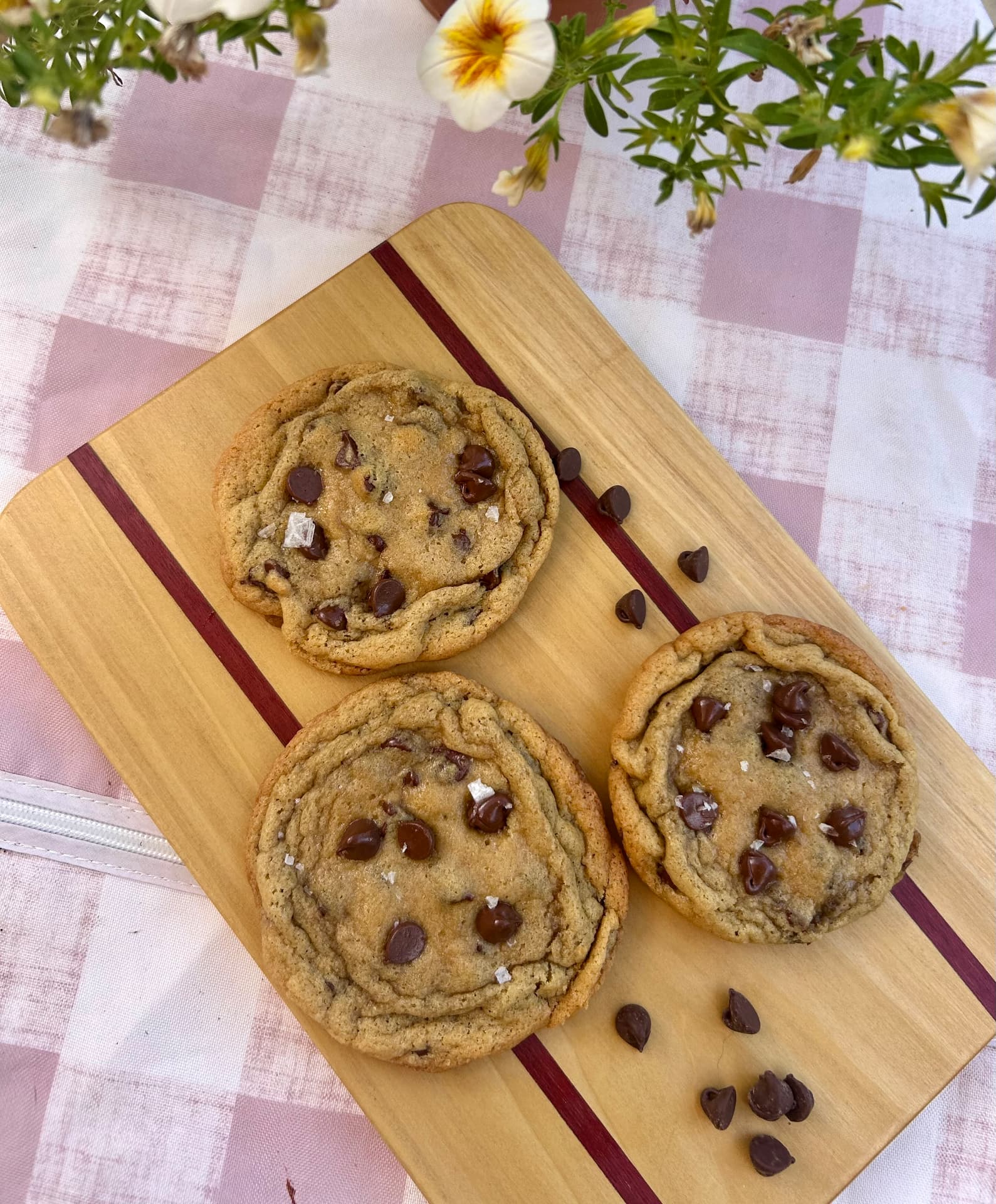 Three big chocolate chip cookies topped with flaky sea salt on a wooden board with scattered chocolate chips
